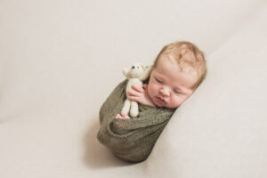 Newborn baby boy in oklahoma city photographed by a photographer while holding a small bear and wrapped in a green wrap