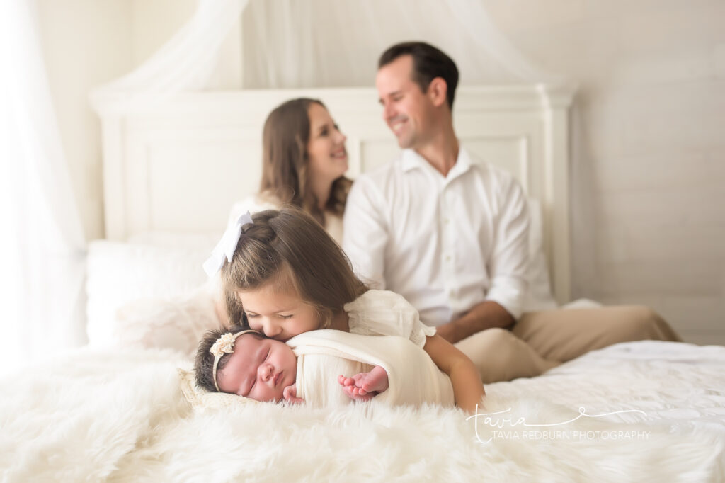 Family sitting on bed at Oklahoma City Newborn Photography session with big sister giving little sister a kiss 